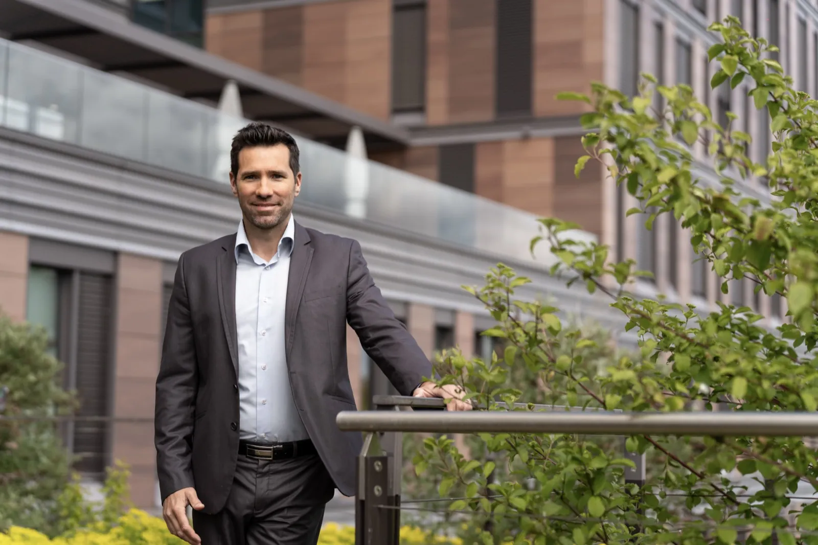 Ein Mann im Anzug steht lächelnd auf einer Terrasse vor einem modernen Gebäude — Frankfurt School